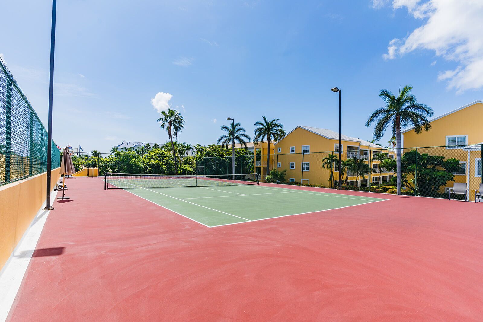 Pista de tenis amb superfície vermella, envoltada per una tanca verda, i al costat d'un edifici groc amb balcons.