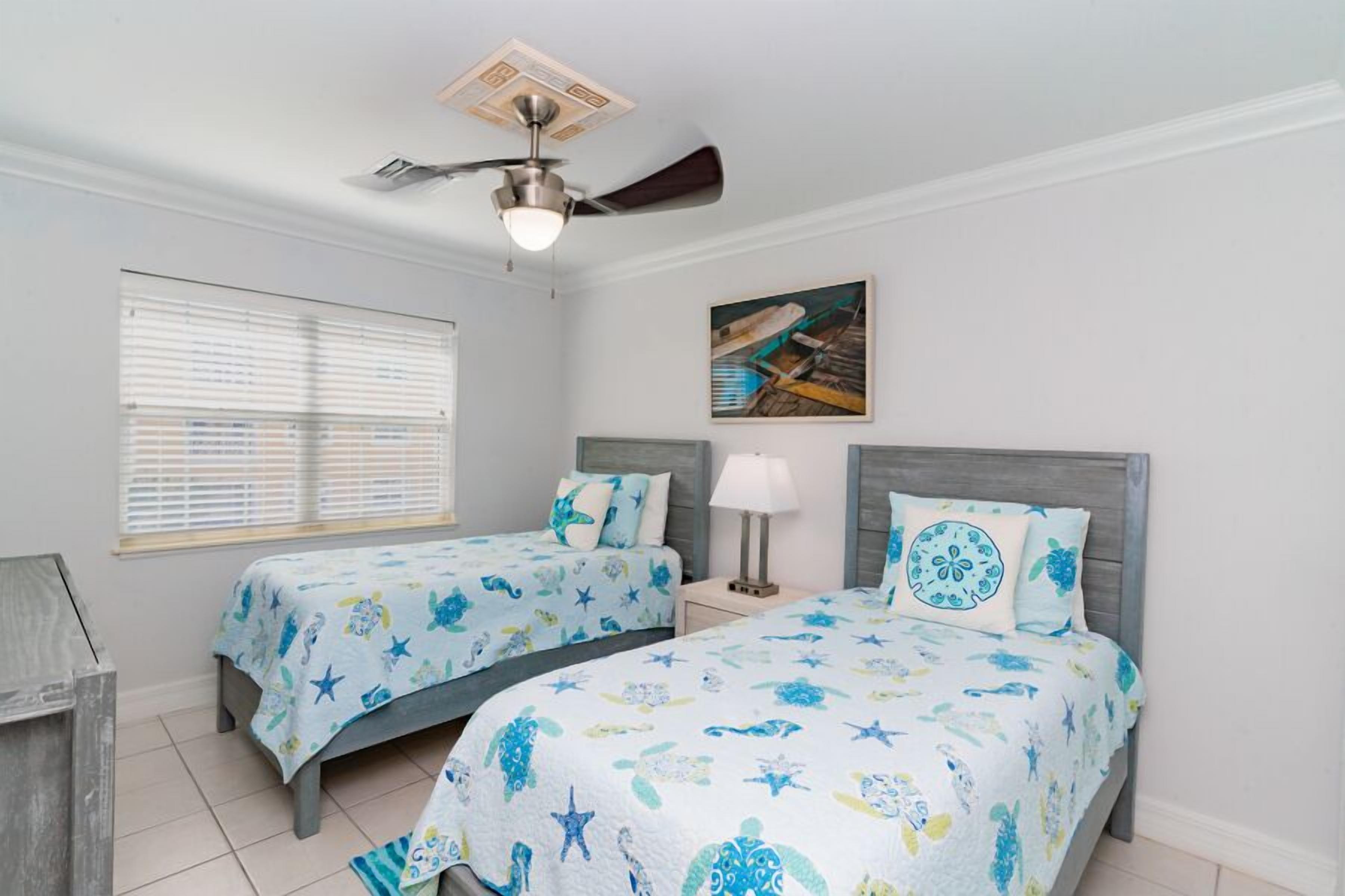 A bedroom with two beds, a ceiling fan, a window with blinds, and a painting on the wall.