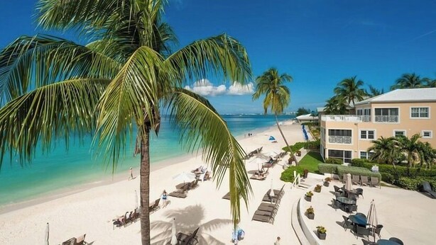 A beach with palm trees, a pool area, and a building with multiple windows. - Regal Beach Club 623 - Oceanfront Condo with Great Views of the Caribbean Sea (Seven Mile Beach)