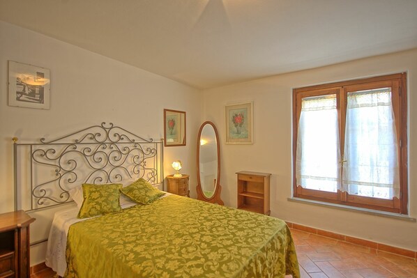 Un dormitorio con estructura de cama metálica, cubrecama verde, una mesita de noche con lámpara, mesita auxiliar de madera, un espejo y una ventana con cortinas.