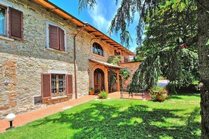 Un edificio de piedra con jardín, un balcón de madera y un gran árbol.