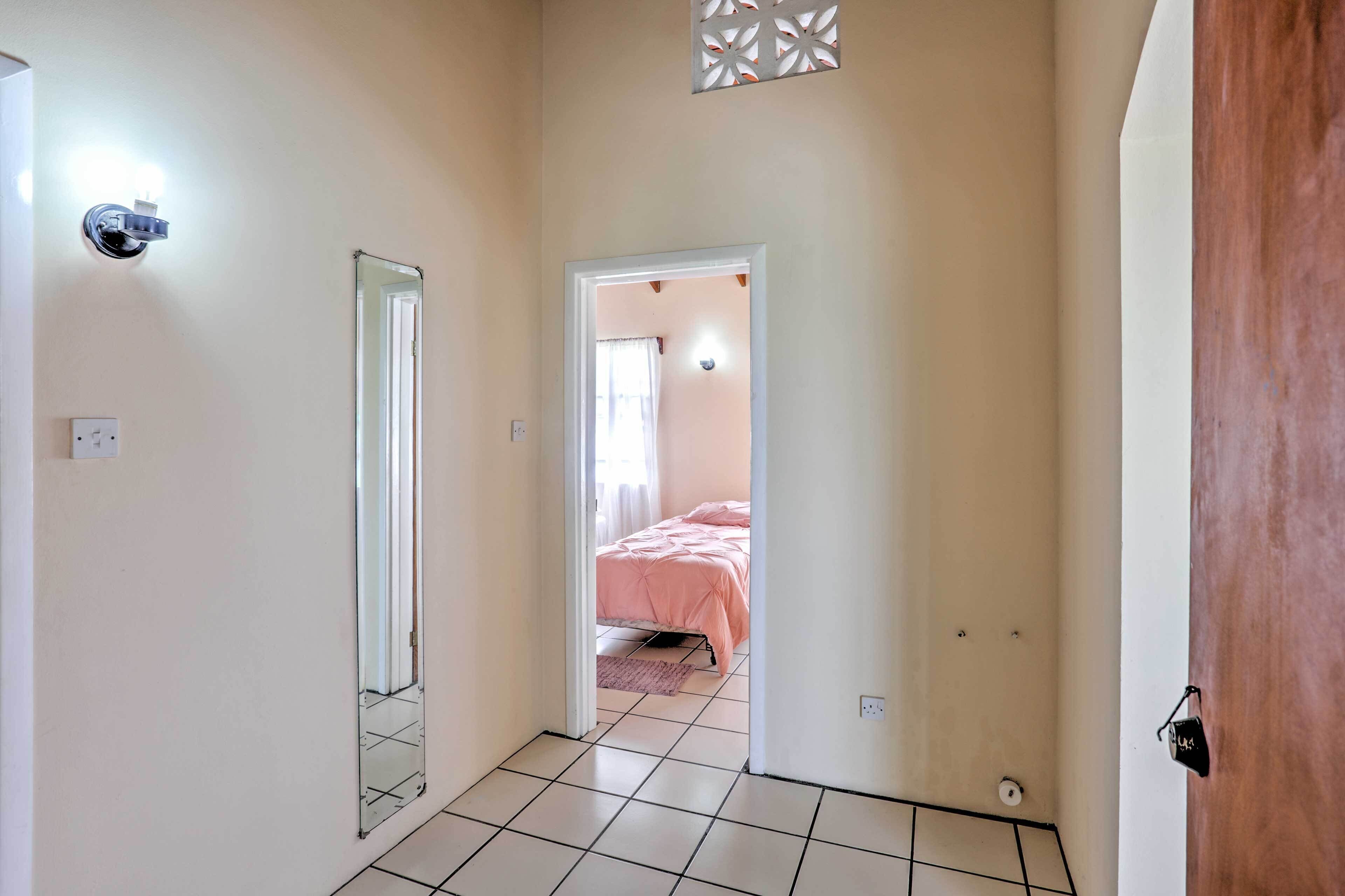 A hallway with a mirror, a door leading to a bedroom with a pink bedspread, and a light fixture on the wall.