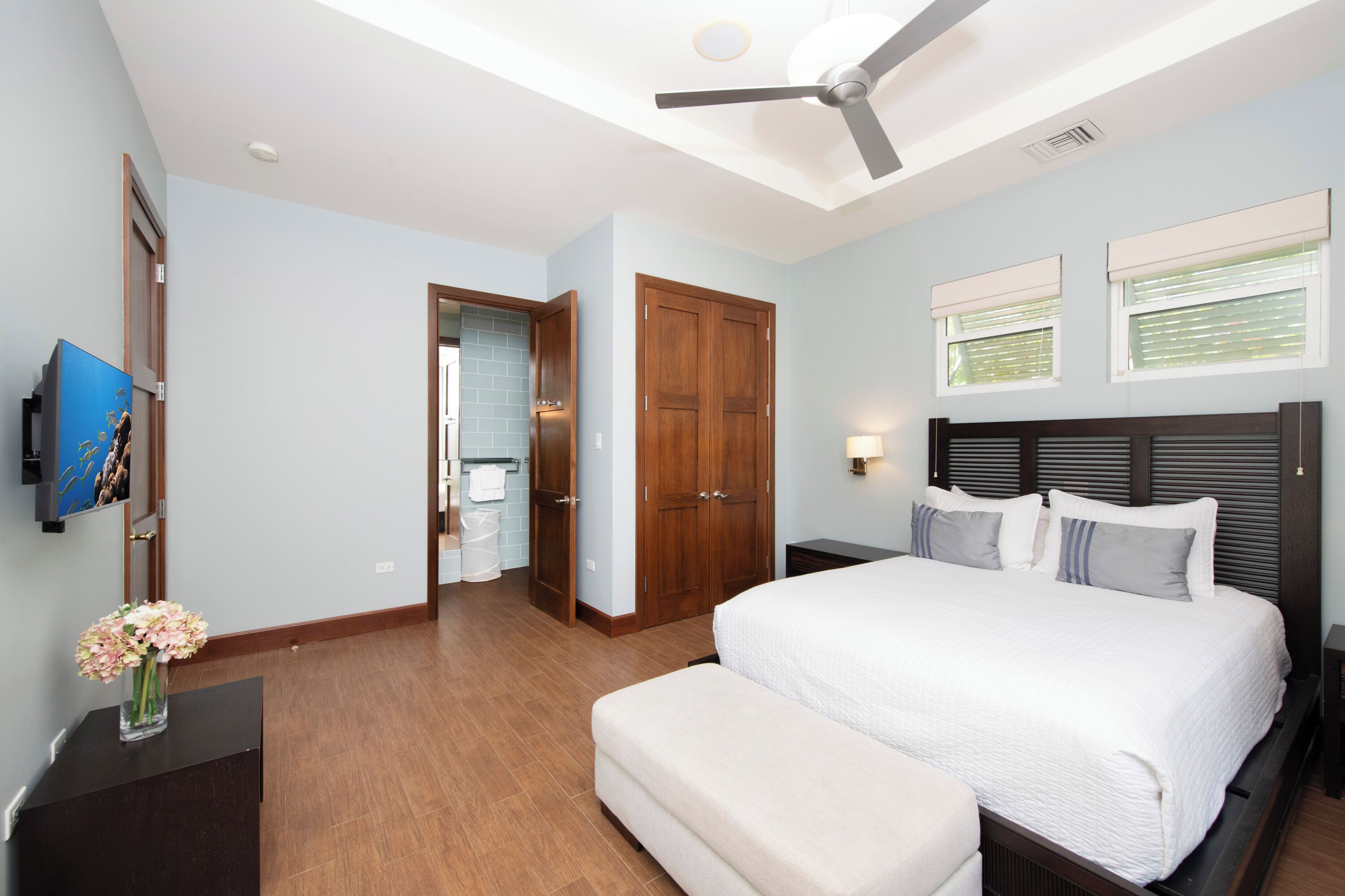 Un dormitorio con cama, mesitas de noche, ventilador de techo y televisor montado en la pared.