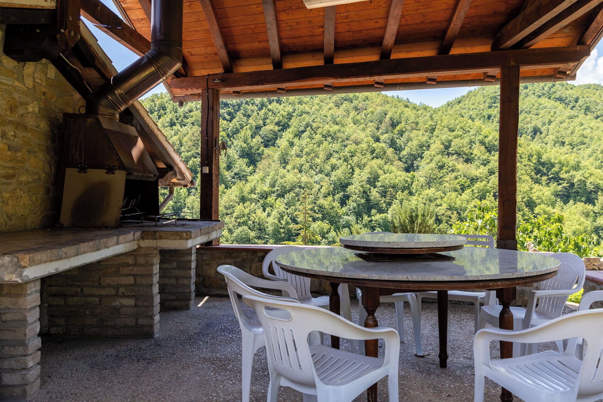 Une terrasse extérieure dotée d’une cheminée en pierre, de chaises en plastique blanches et offrant une vue sur une montagne boisée.