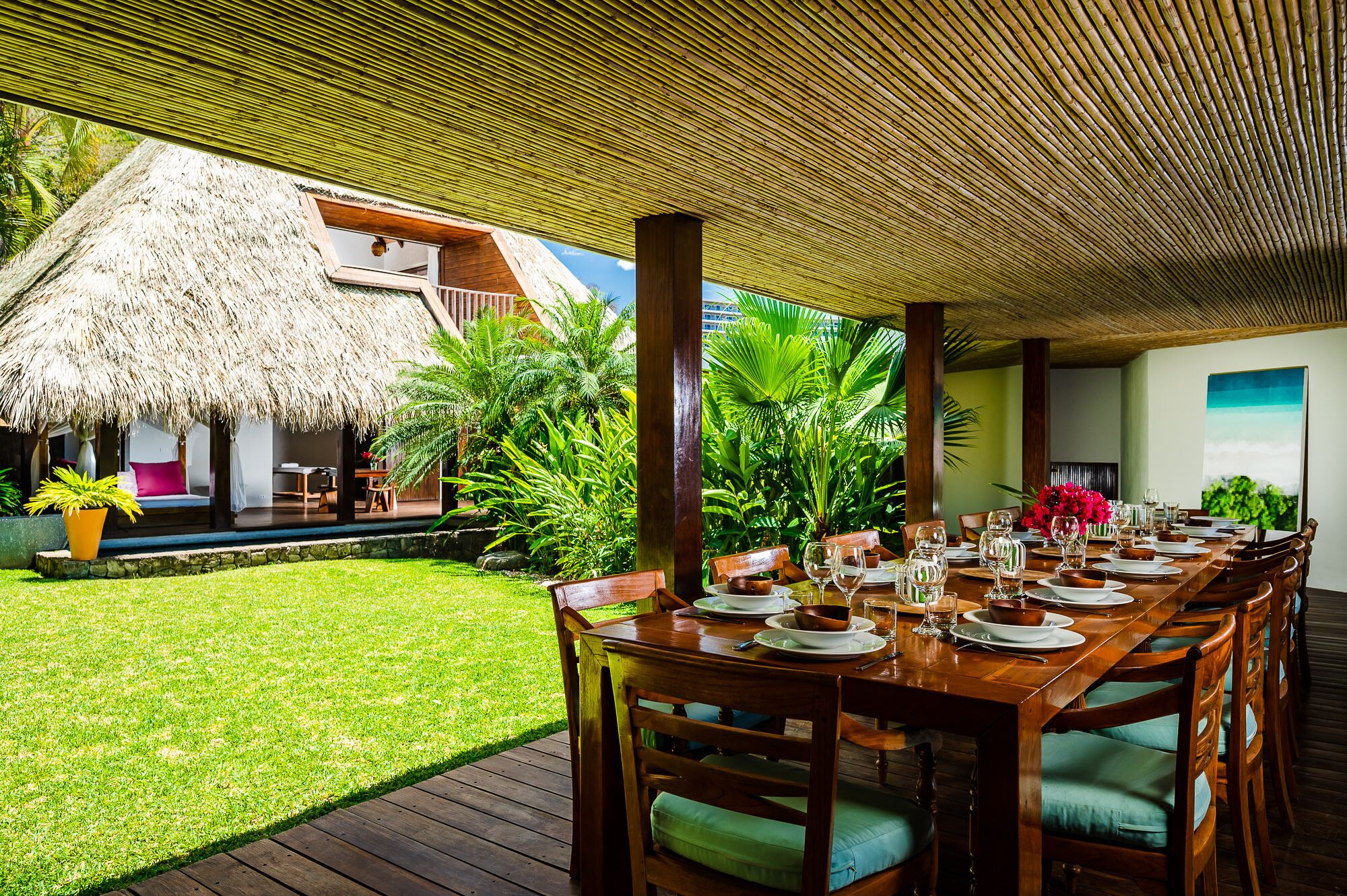 An outdoor dining area with a thatched-roof structure, wooden tables, and lush greenery.