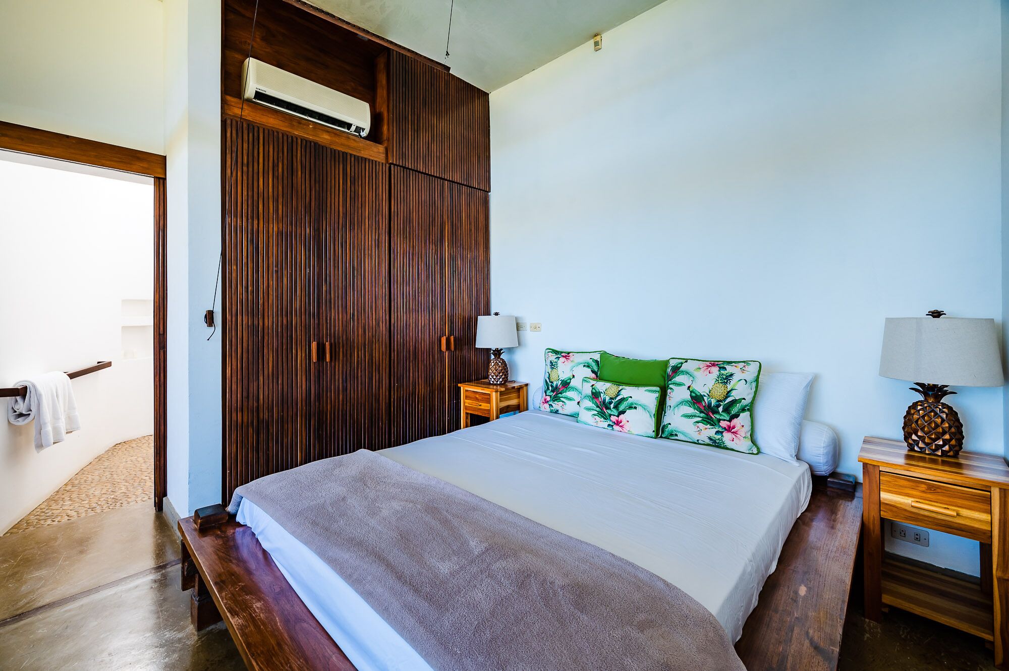A hotel room with a large bed, wooden wardrobe, two bedside tables with lamps, and a tropical-themed pillow.