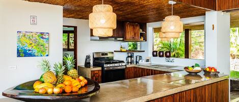 A modern kitchen with a wooden ceiling, a large island with fruit, and a view of greenery outside.
