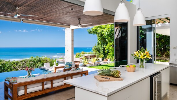 A modern kitchen with a large island, a dining area with a wooden bench, and a view of the ocean. - Ocean Views · Infinity Pool · 3 min to the Beach & Exclusive Beach Club Access (Tamarindo)