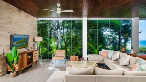 A modern living room with a wooden ceiling, large windows, a sofa, a coffee table, and a television. - Ocean Views · Infinity Pool · 3 min to the Beach & Exclusive Beach Club Access (Tamarindo)