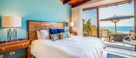 Une chambre avec un grand lit, une tête de lit en bois, des lampes de chevet et une vue sur l’océan, visible à travers une porte-fenêtre coulissante.