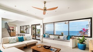 A spacious living room with a large window offering a view of the sea, a ceiling fan, and a modern sofa set.