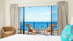 A bedroom with a large bed, two rocking chairs, a small table with drinks, and a view of the sea through floor-to-ceiling windows.