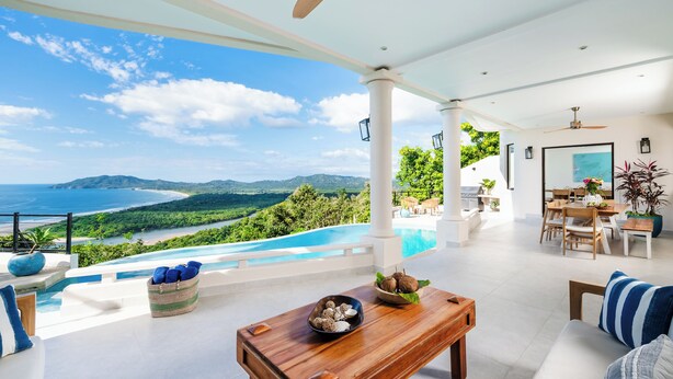 A spacious outdoor area with a pool, wooden furniture, and a scenic view of the ocean and mountains.
