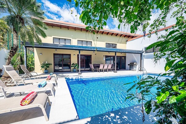 Un espace aménagé au bord de la piscine, avec des chaises longues et un coin salon abrité, le tout entouré d’une végétation luxuriante et d’un bâtiment doté de grandes fenêtres.