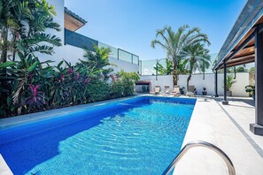 Une piscine dont les bords sont recouverts de carreaux, entourée d’une végétation tropicale luxuriante et bordée d’un bâtiment moderne doté d’un balcon.