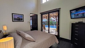 A bedroom with a bed, a TV, a desk with a lamp, a window with blinds, and a door. - Casa Gris with private pool near the beach in Jaco (Jacó)