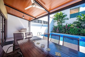 Un coin repas avec une table et des chaises en bois, un ventilateur de plafond et une vue sur une piscine.