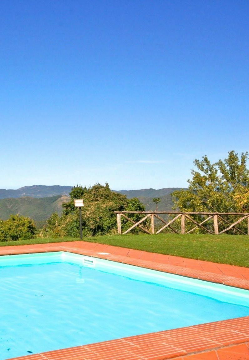 A rectangular swimming pool with a clear blue water surface, surrounded by a red brick deck. In the background, there is a wooden fence and a view of distant mountains under a clear blue sky.