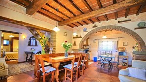 A spacious living area with a wooden dining table, chairs, and a sofa. There is a kitchen area with a stove and a microwave. The room features exposed wooden beams and a tiled floor. - Villa Margherita Uno - Holiday Villa Rental with swimming pool in Cortona, Tuscany (Cortona)