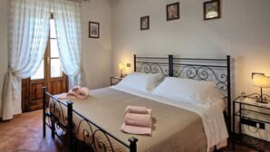 A neatly made bed with a metal headboard, a bedside table with a lamp, and a window with sheer curtains. - Villa Portole Uno - Holiday Villa Rental in Cortona (Tornia)