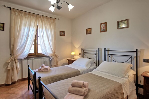 A room with two beds, a window with curtains, a radiator, and a wall with framed pictures. - Villa Portole Uno by PosarelliVillas (Tornia)