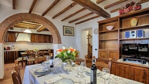 A dining area with a table set for four, a brick archway, wooden beams, and a kitchen in the background. - Villa Portole Uno - Holiday Villa Rental in Cortona (Tornia)