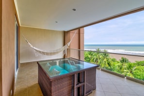 Uma varanda com mesa de vime e uma rede, com vista para uma praia e palmeiras.