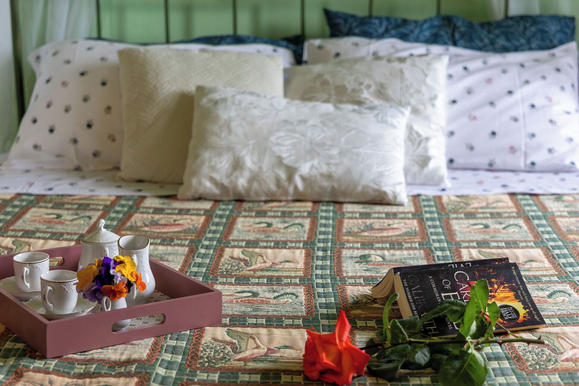 Ein Bett mit gemusterten Bettwaren, ein Tablett mit Tassen und Blumen sowie ein Buch auf dem Bett.