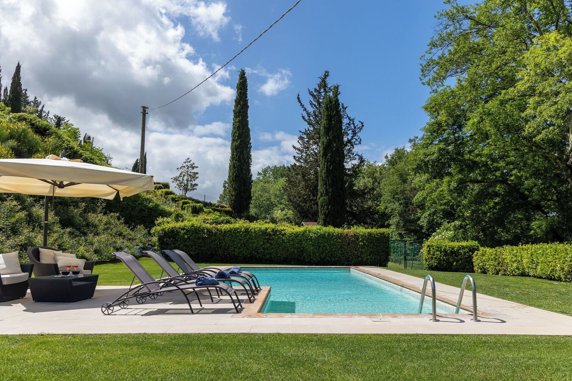 A swimming pool with lounge chairs and a sun umbrella, surrounded by well-maintained hedges and tall trees.