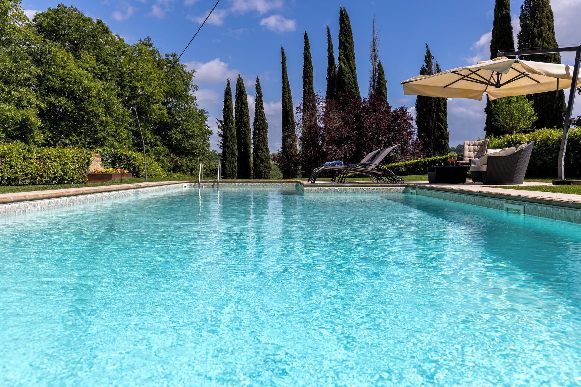 A clear swimming pool with lounge chairs and a sun umbrella, surrounded by tall trees and a well-maintained hedge.
