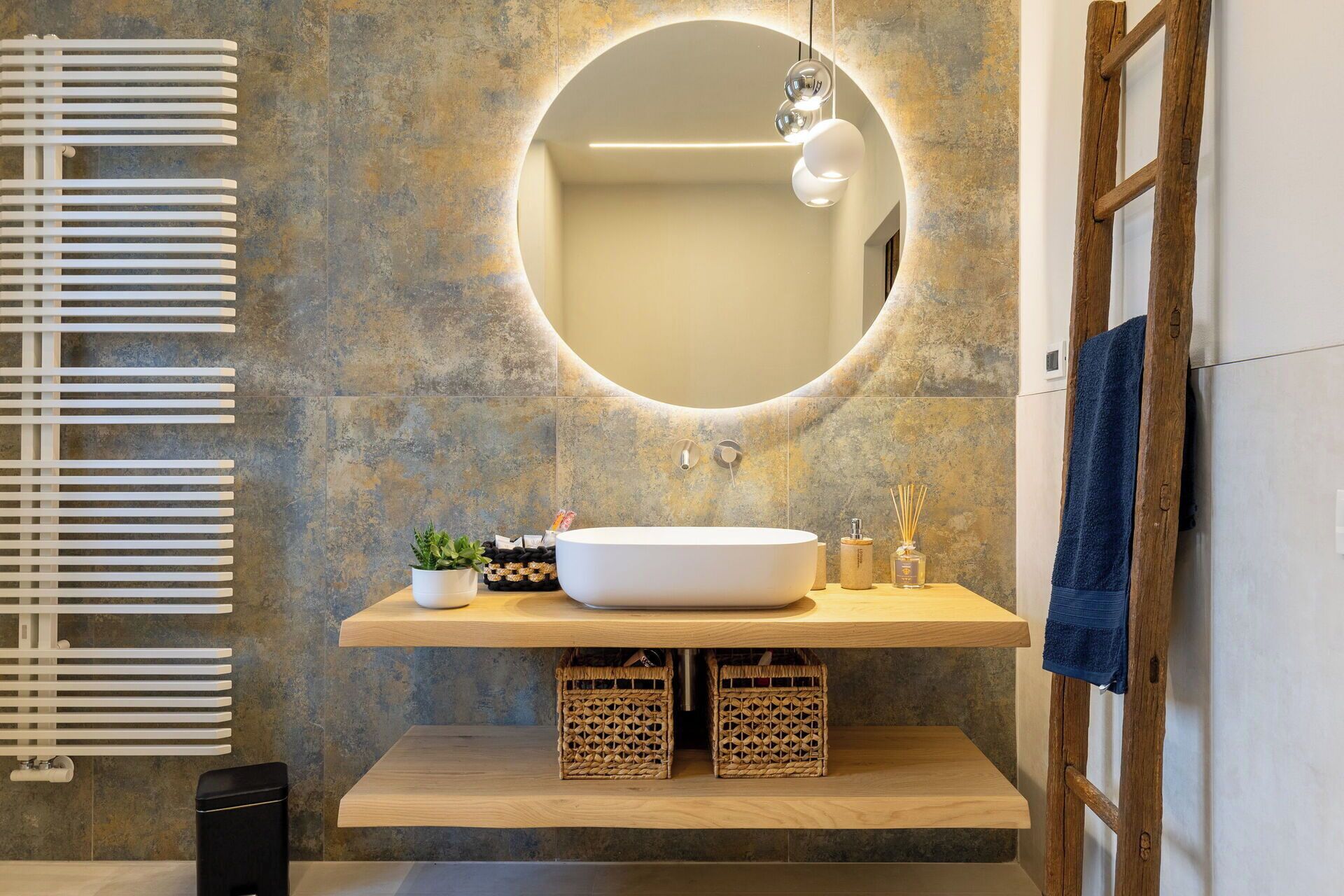 A modern bathroom with a large oval mirror, a white sink, wooden shelves, and a towel rack.