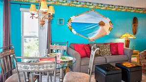 Ein Zimmer mit einer türkisblauen Wand, einem Strandgemälde, einem Esstisch mit Stühlen, einer Couch mit roten Kissen und einer Hängelampe.