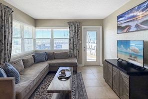 A living room with a sectional sofa, a coffee table, a TV on a cabinet, and large windows with curtains.