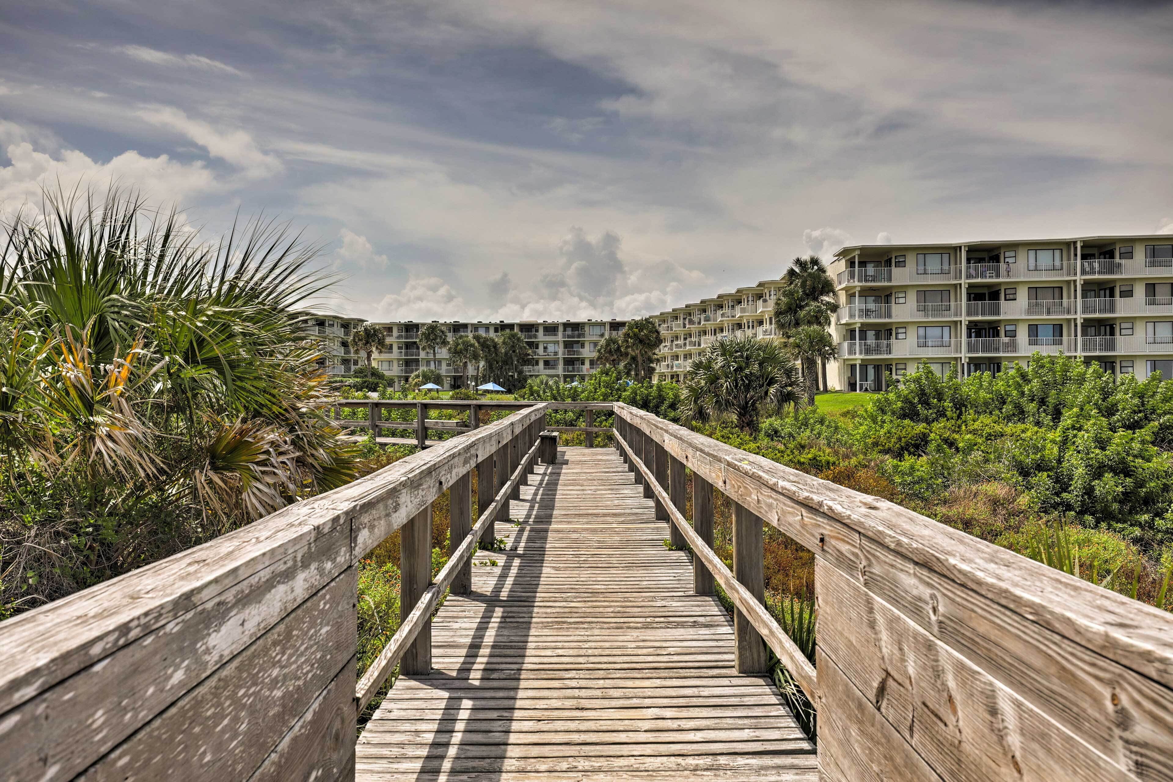 A wooden boardwalk leading to a multi-story building with balconies, surrounded by greenery.