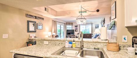 A kitchen with granite countertops, stainless steel sink, and a dishwasher. There is a chandelier above the sink and a sign on the wall that reads "Beach House."