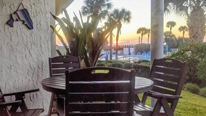 Eine Terrasse mit rundem Tisch und vier Stühlen, mit Blick auf einen Poolbereich mit Palmen und Sonnenuntergang.