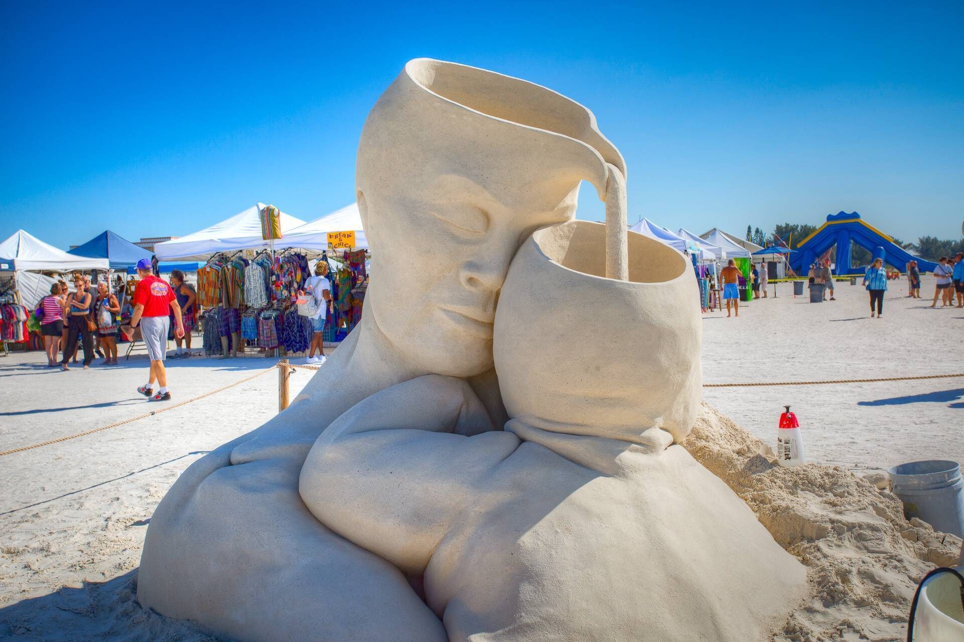 A sand sculpture of a face with a cup, located on a beach with tents and people in the background.
