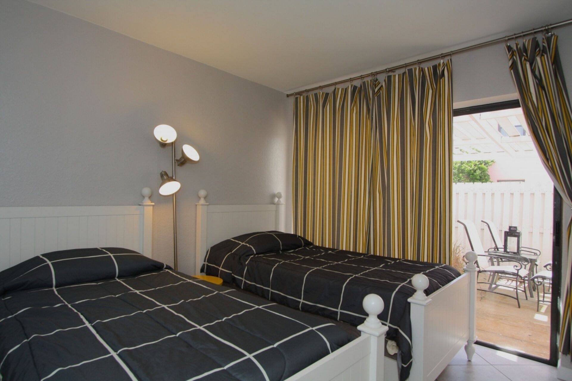 A bedroom with two beds, a bedside table with lamps, a window with curtains, and a view of the outside patio.