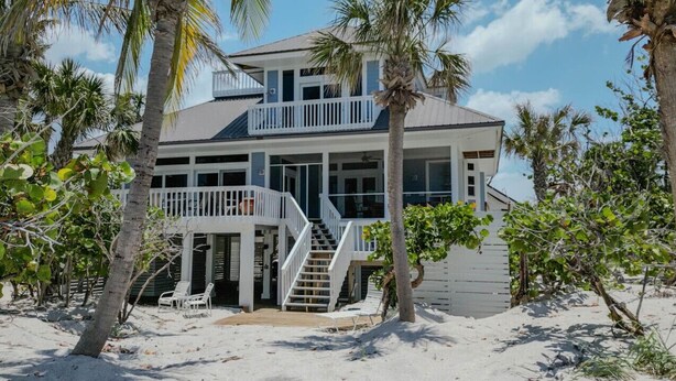 A beachfront house with a balcony, a staircase leading to the upper floor, and a sandy beach area with lounge chairs. - Manatee Manor: Exclusive Beachfront Retreat with Gulf Views (North Captiva)