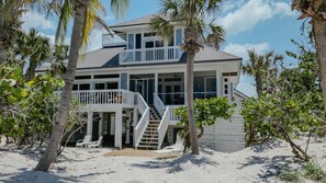 Una casa frente a la playa con balcón, una escalera que lleva al piso superior y un área de playa con sillas de descanso.