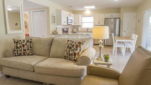 A living room with a beige sofa, a coffee table, and a lamp. There is a kitchen in the background with white cabinets and a refrigerator. - Beach Villa ~ Pet Friendly ~ Fenced Yard ~ Steps to the Beach!!! (Saint Augustine)