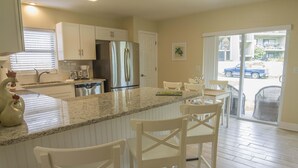 A modern kitchen with white cabinetry, a stainless steel refrigerator, a granite countertop, and a dining area with white chairs. - Beach Villa ~ Pet Friendly ~ Fenced Yard ~ Steps to the Beach!!! (Saint Augustine)