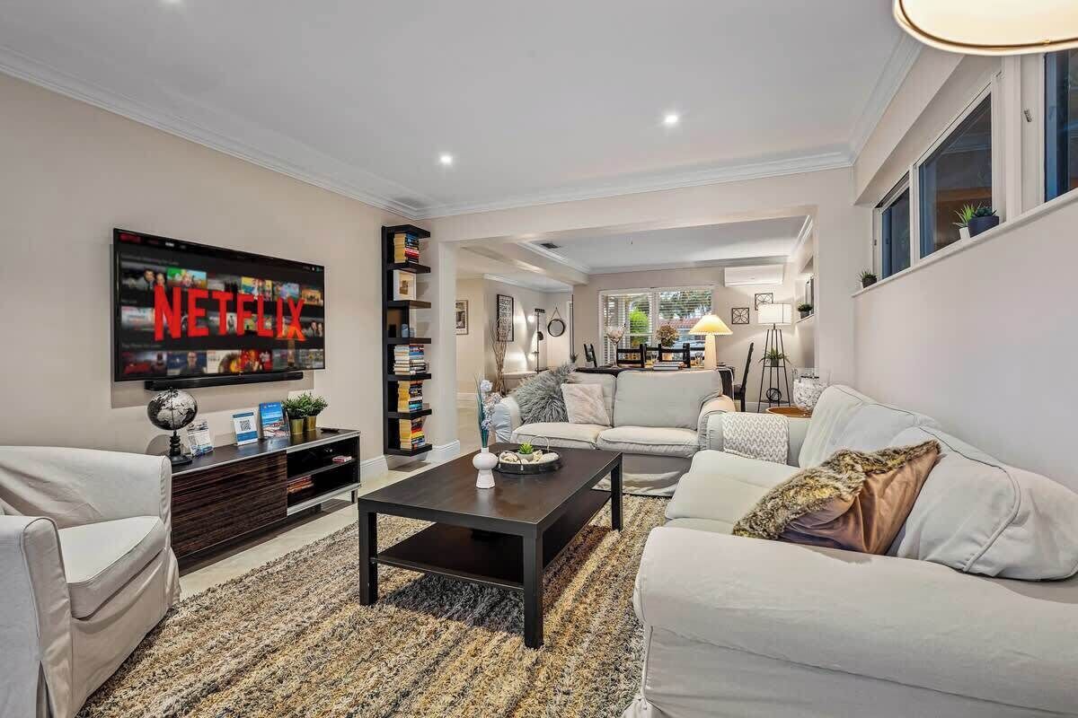 A modern living room with a large flat-screen TV displaying the Netflix logo, a dark coffee table, a white sofa, and a bookshelf.