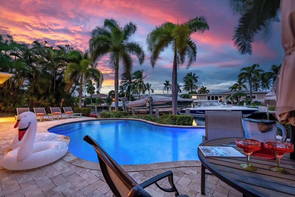A swimming pool with lounge chairs and a swan float, surrounded by palm trees and a sunset sky.