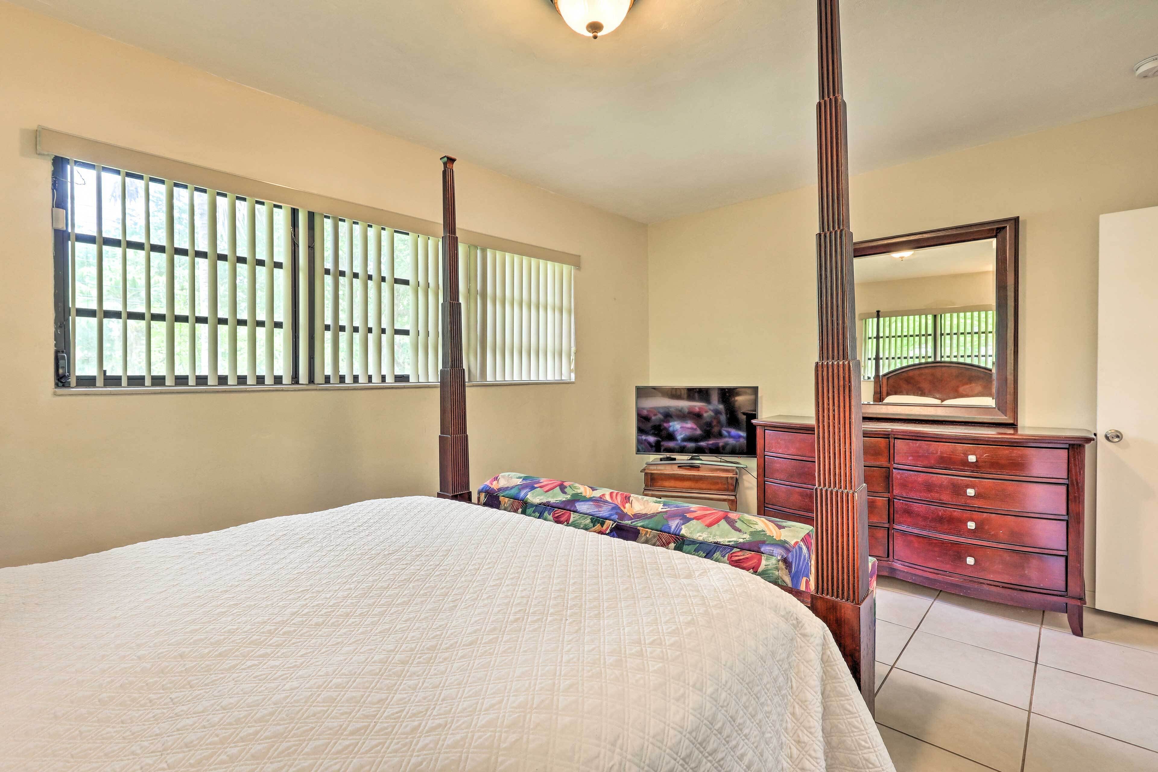 A bedroom with a large bed, a dresser, a television, and a window with blinds.
