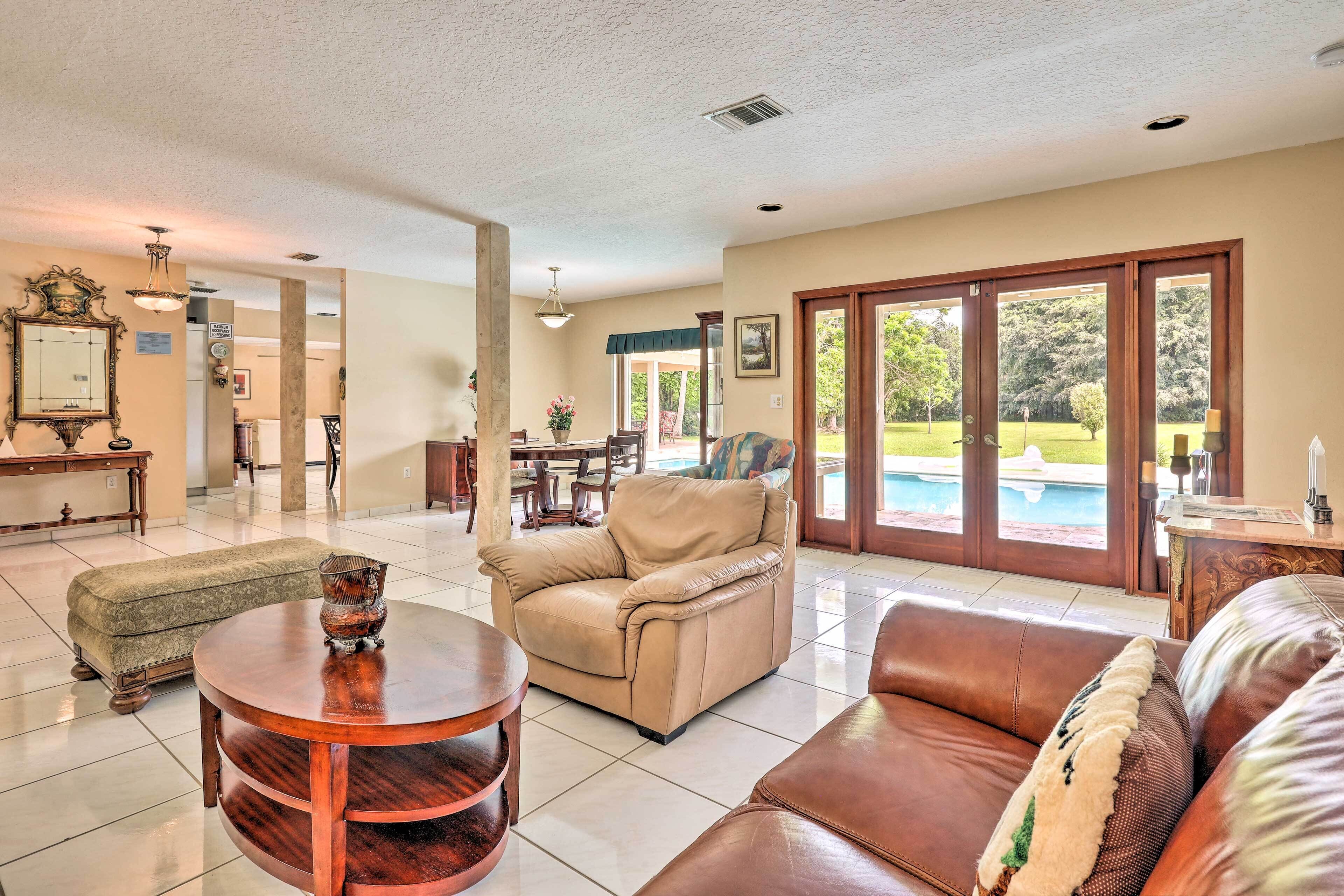 A spacious living room with a beige sofa, a brown leather couch, and a round wooden coffee table. There is a dining area with a table and chairs, and a large window offering a view of a swimming pool.