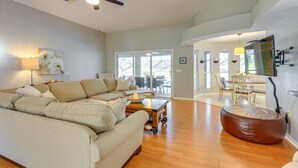 A spacious living room with a large beige sectional sofa, a wooden coffee table, and a brown ottoman. A ceiling fan and a TV mounted on the wall are visible. - 3 Mi to Dunnellon Historic District: Home w/ Lanai (Dunnellon)