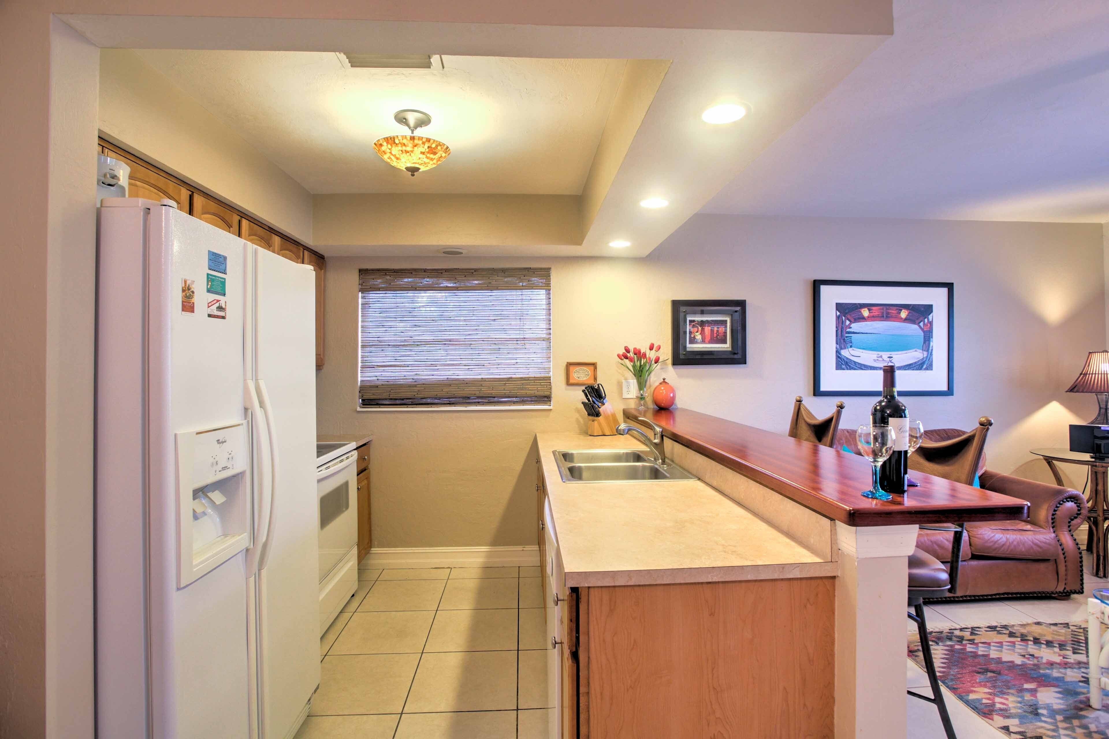 A compact kitchen with a refrigerator, sink, and wooden counter. There is a window with blinds, a framed picture on the wall, and a small table with chairs.