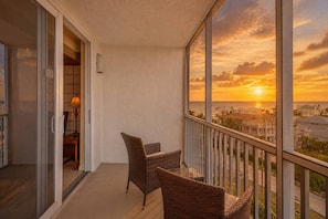 A balcony with wicker chairs, a glass table, and a view of the sunset over the ocean.
