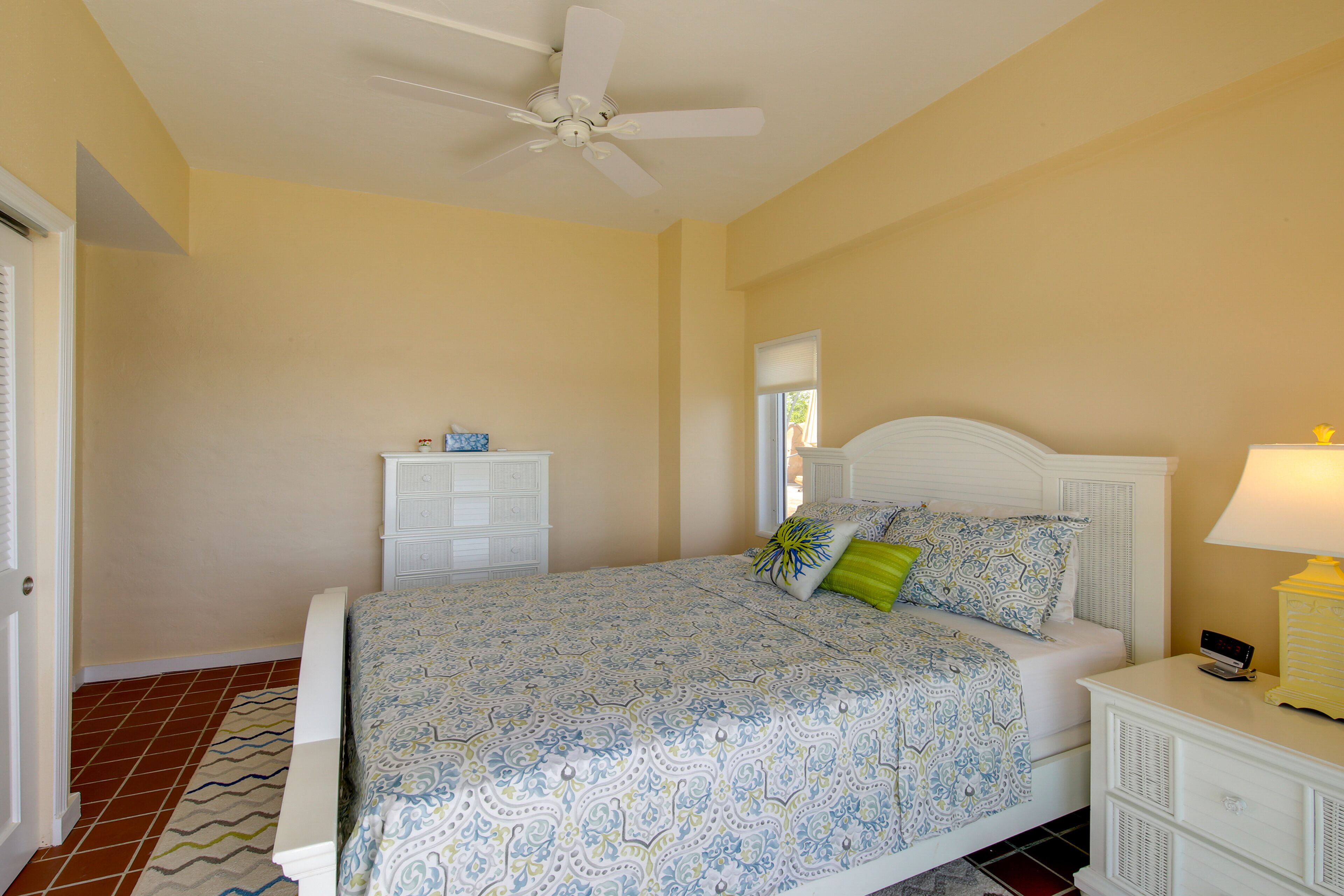 Quarto com cama, mesa de cabeceira, cômoda, ventilador de teto e janela.